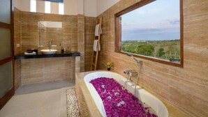 A modern bathroom with a large bathtub filled with pink flowers, a sink, and a window offering a view of greenery. - Kandahill Bali (Pecatu)