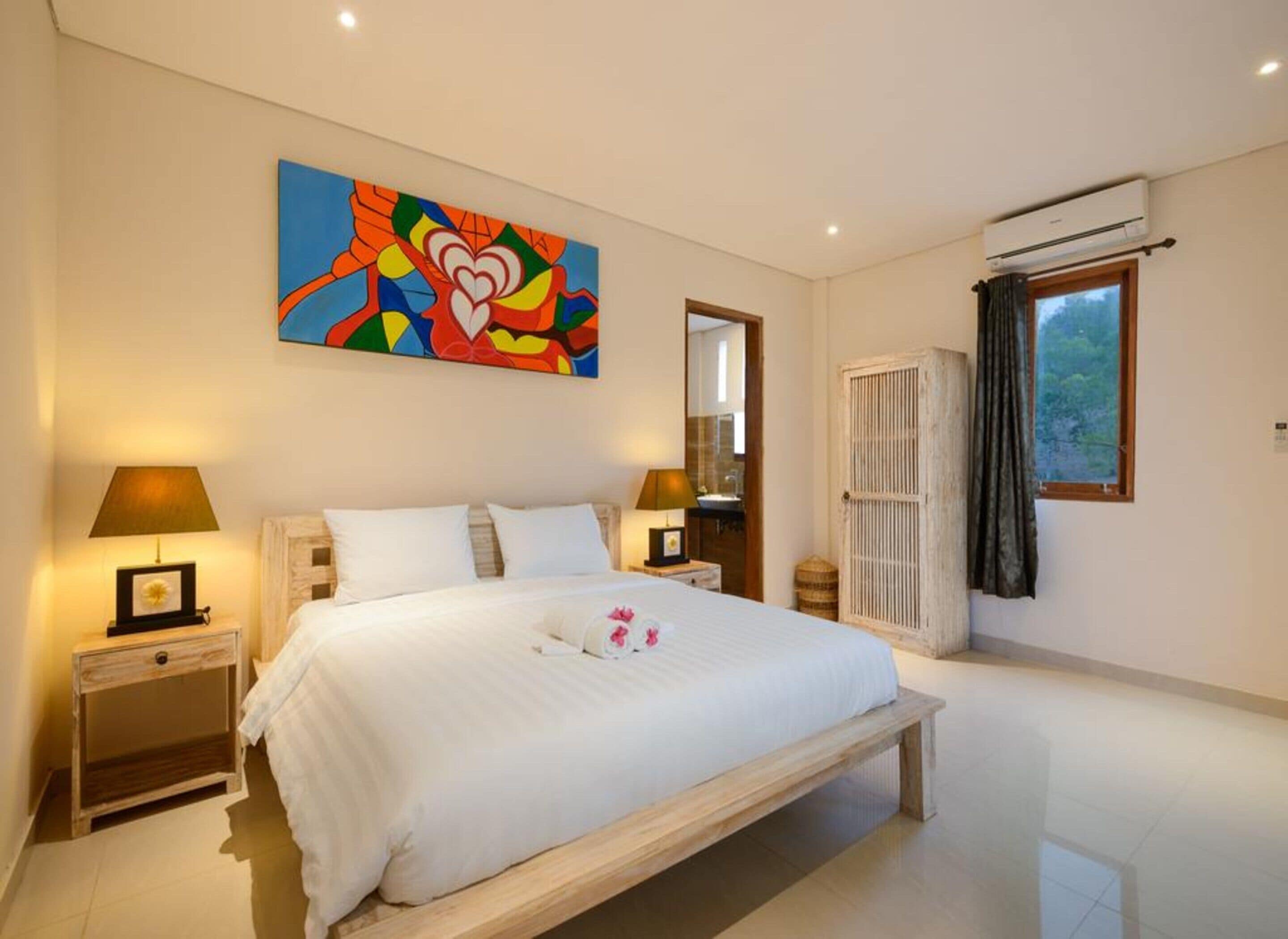 A bedroom with a large bed, two bedside tables with lamps, a colorful abstract painting, a window with curtains, and an air conditioning unit.