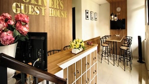 A hotel lobby with a wooden reception desk, a coffee machine, a flower vase, and a dining area with chairs and a table. - Oliver's Guest House (Pagedangan)