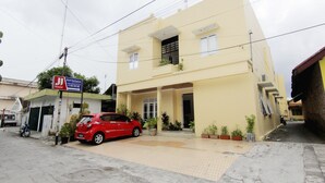 Un edificio a due piani con un'auto rossa parcheggiata davanti. Vasi con piante adornano l'ingresso e un cartello indica un'abitazione.