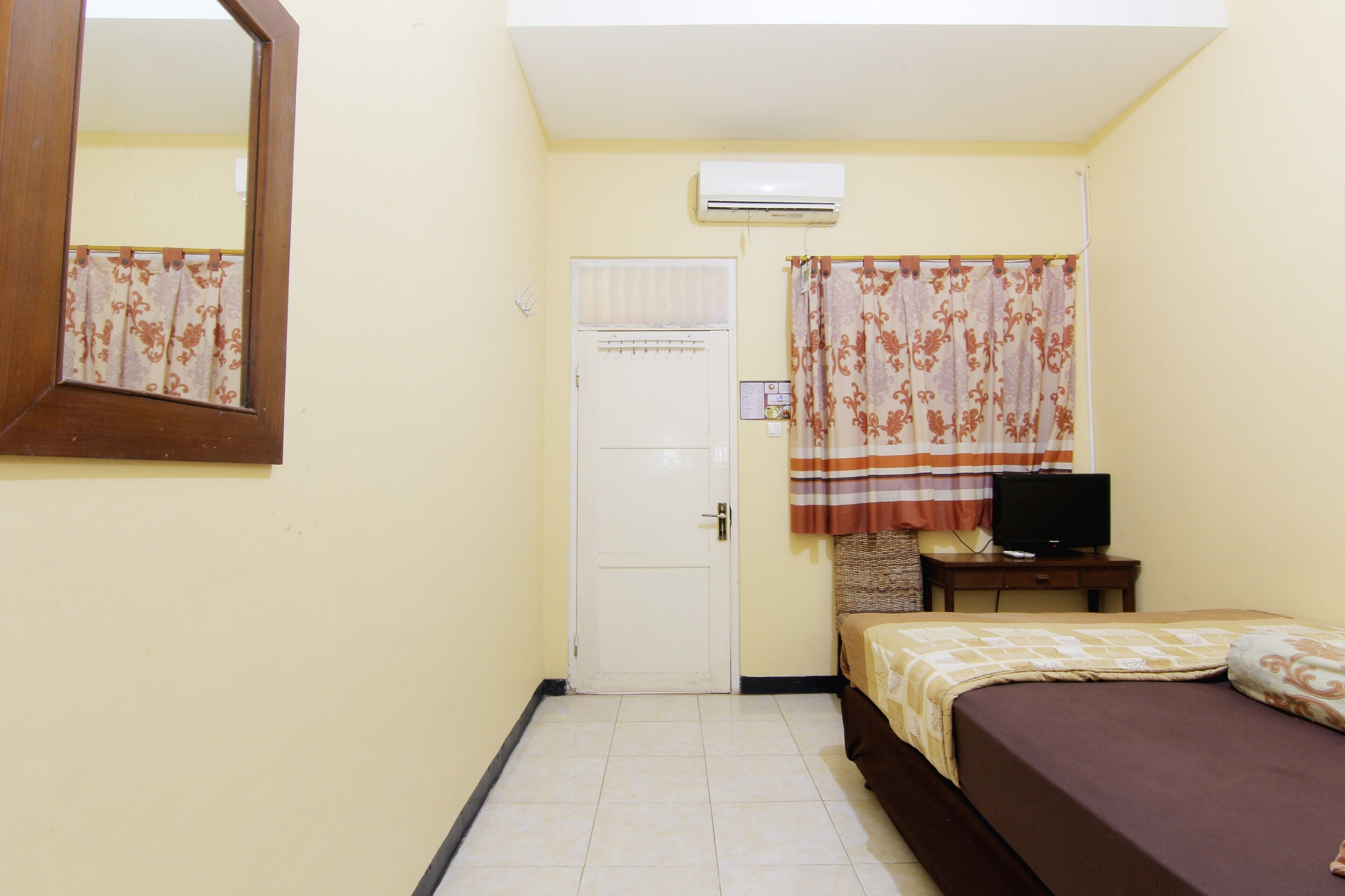 Una habitación pequeña y limpia con cama, televisión sobre una mesa de madera, espejo y puerta.