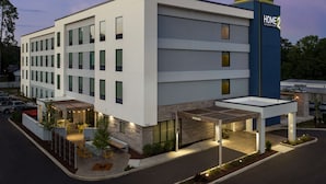 Un hôtel moderne à la façade sobre, doté de nombreuses fenêtres et d’un espace extérieur aménagé avec des sièges, protégé par un auvent.