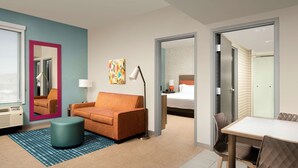 Une chambre d’hôtel avec un canapé, un pouf, une table et des chaises. Une chambre adjacente contient un lit.