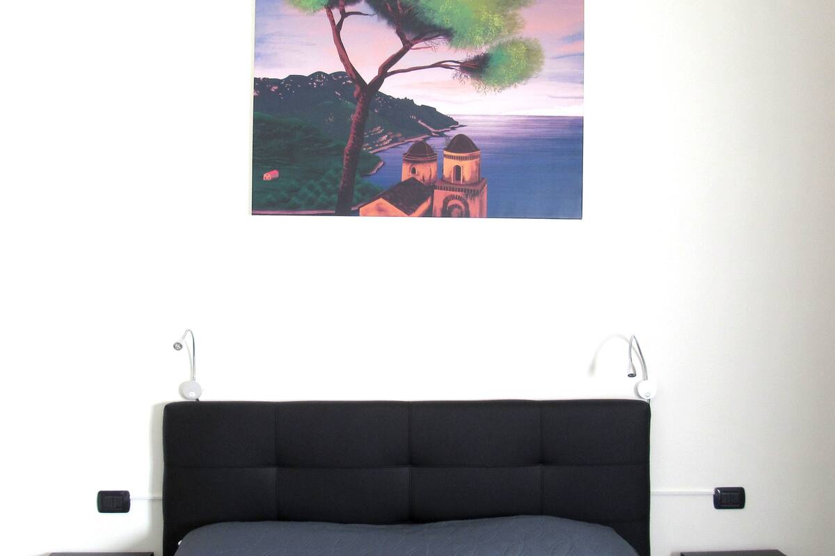 A bedroom with a large bed, a nightstand, a wall-mounted lamp, and a painting of a landscape.