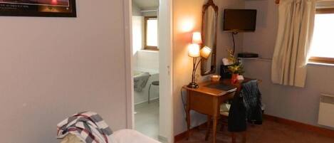 A hotel room with a bed, a desk with a lamp, a mirror, a television, and a window with curtains.