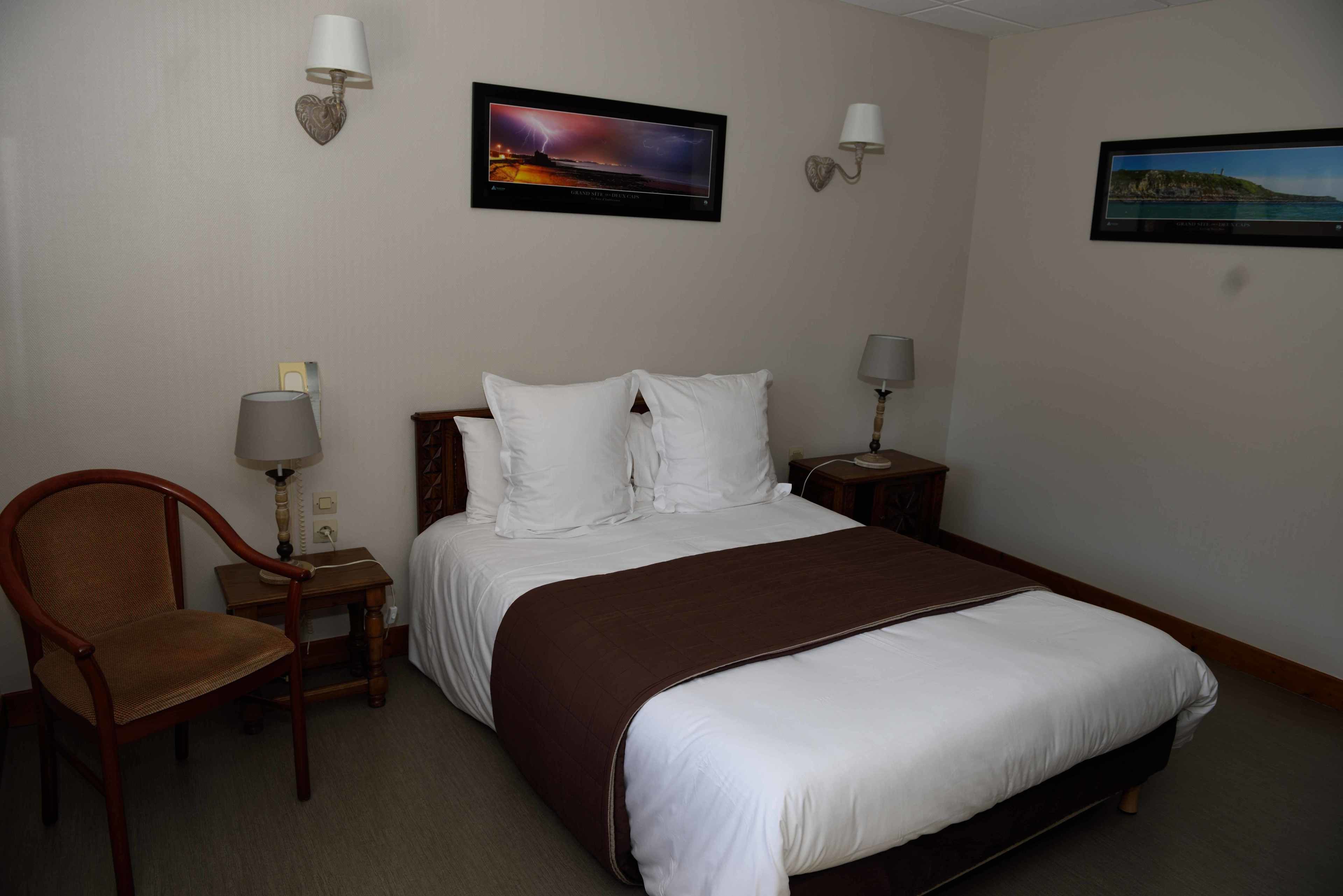 Habitación de hotel con una cama, una silla, dos mesitas de noche con lámparas y dos cuadros colgados en la pared.