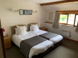 Habitación doble con cama, cabecera, mesitas de noche, lámpara de pared, ventana con cortinas y radiador.