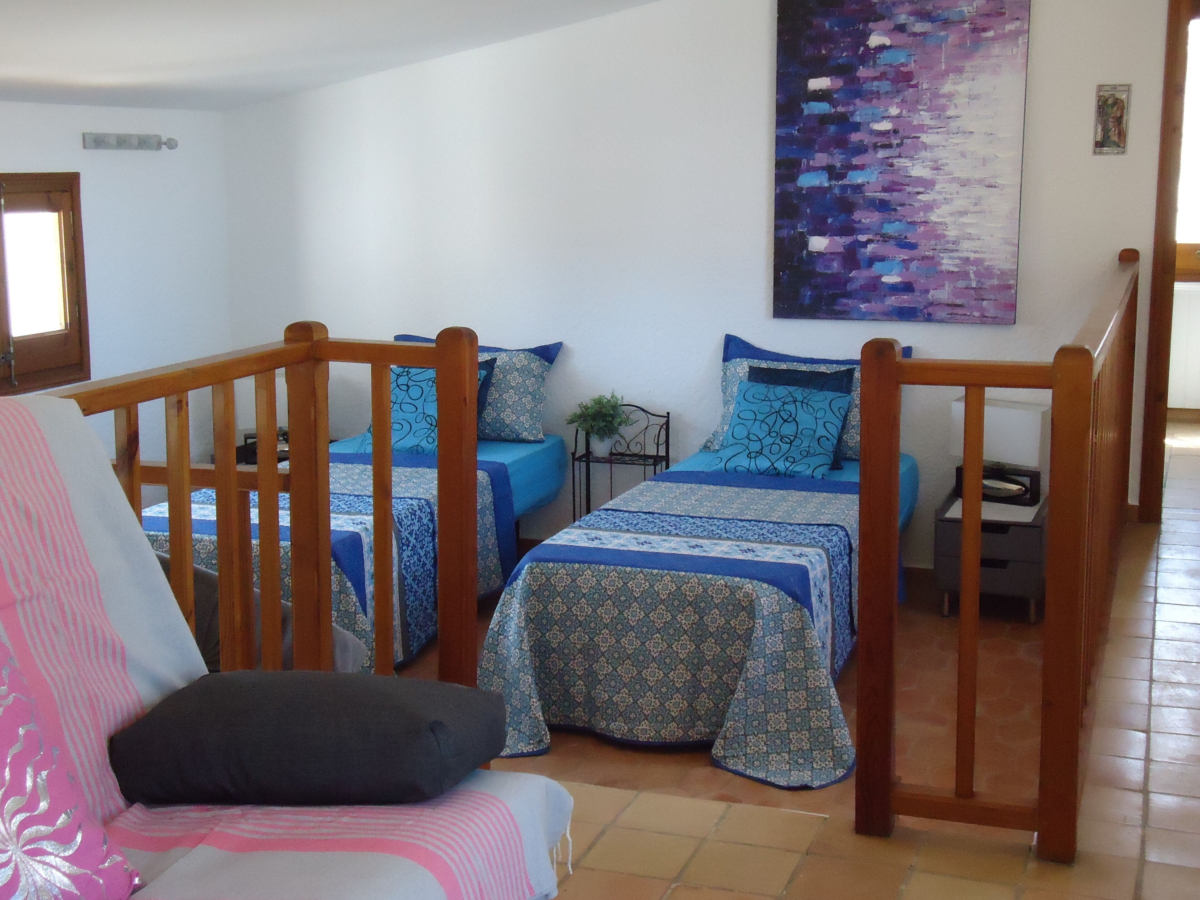 A room with two beds, each with a wooden frame and blue bedding. There is a chair with a pink and white striped cushion. A painting is on the wall above the beds.