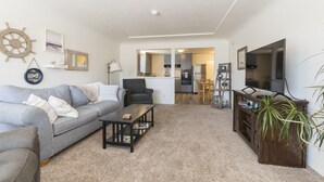 A living room with a sofa, coffee table, and TV stand. There is a kitchen in the background with cabinets and a refrigerator. - Beach cottages, majestic Morro Bay (Morro Bay)