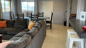 A living room with a grey sectional sofa, a dining table with chairs, and a kitchen area with a bar counter and stools. - Large Casa in Rocky Point, Mexico (Puerto Peñasco)