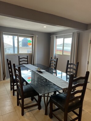 A dining area with a rectangular glass table surrounded by wooden chairs, large windows with curtains, and a view of a building outside.
