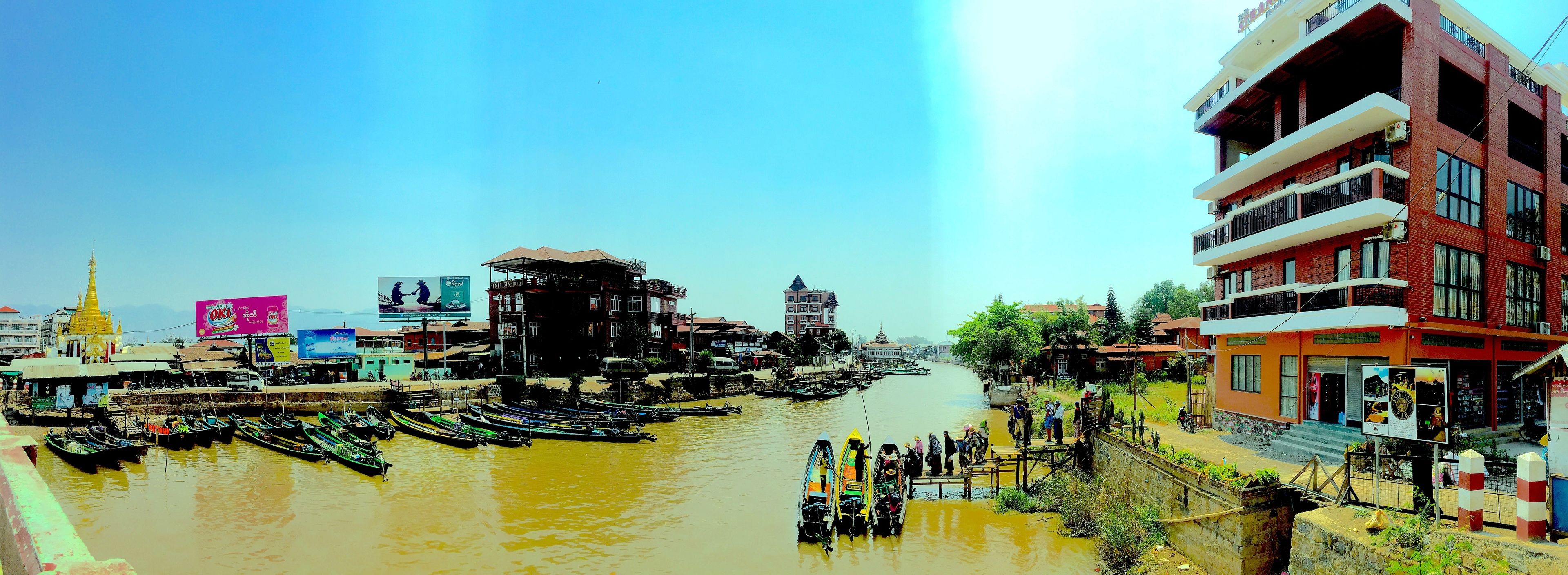 A riverside area with boats docked along the water, buildings with balconies, and a clear sky.
