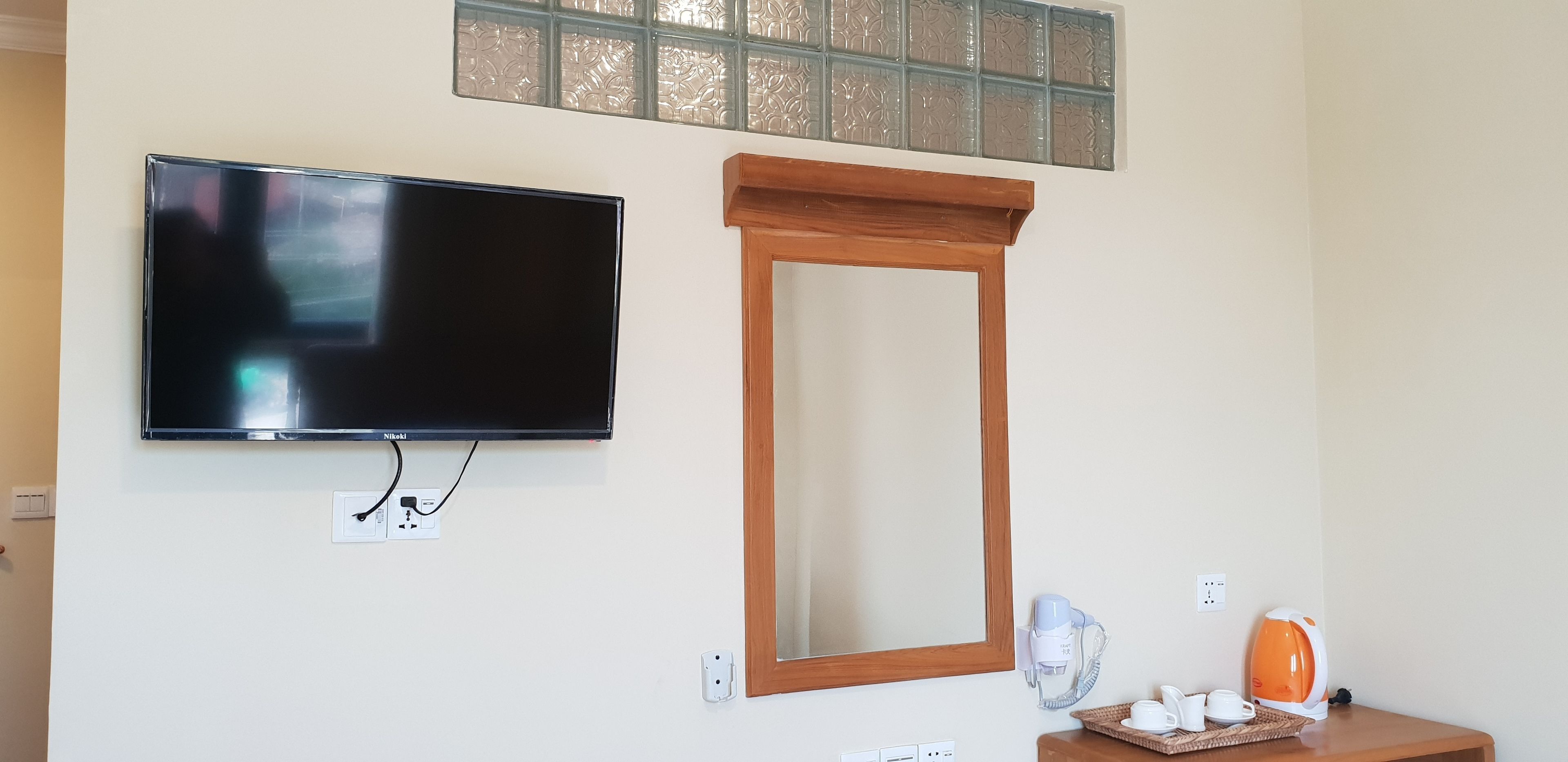 Une chambre dotée d’un téléviseur à écran plat fixé au mur, d’un miroir encadré en bois et d’une petite table sur laquelle se trouvent une bouilloire et un sèche-cheveux.