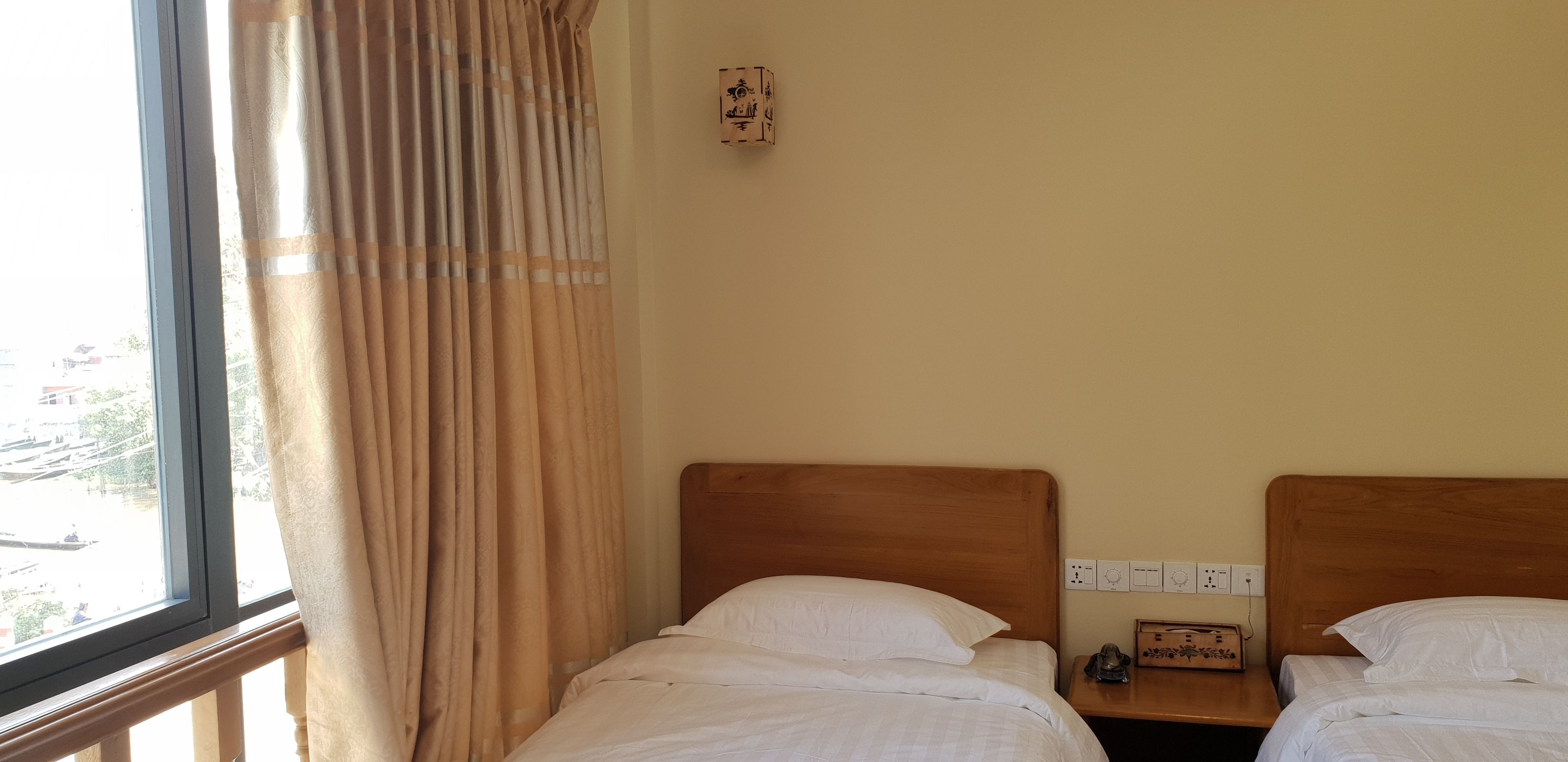 A hotel room with two single beds, a window with curtains, a bedside table with a lamp, and a wall socket.