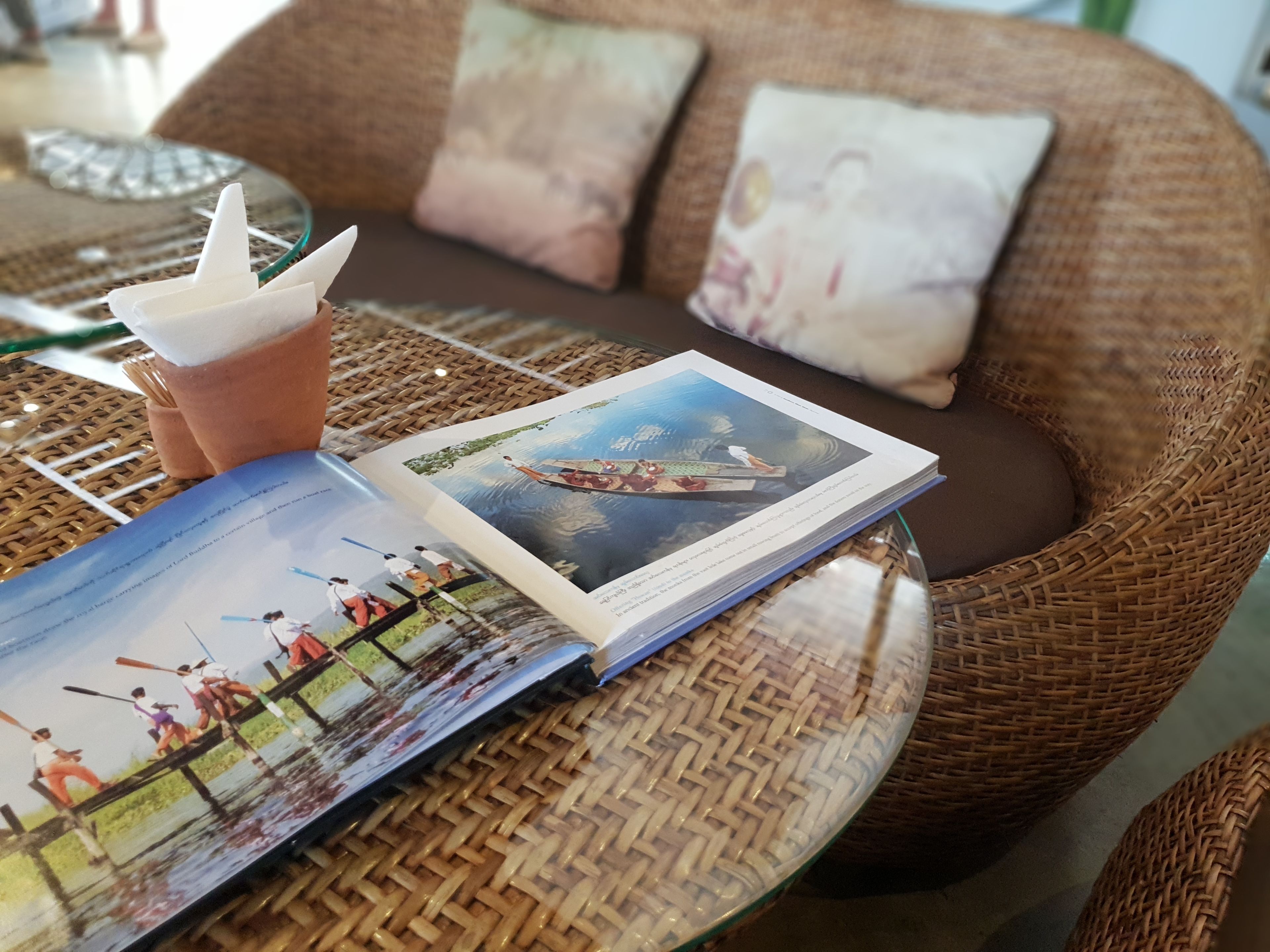Una silla de mimbre con un cojín, una mesa de cristal con una revista y una bebida, y una cesta de mimbre.