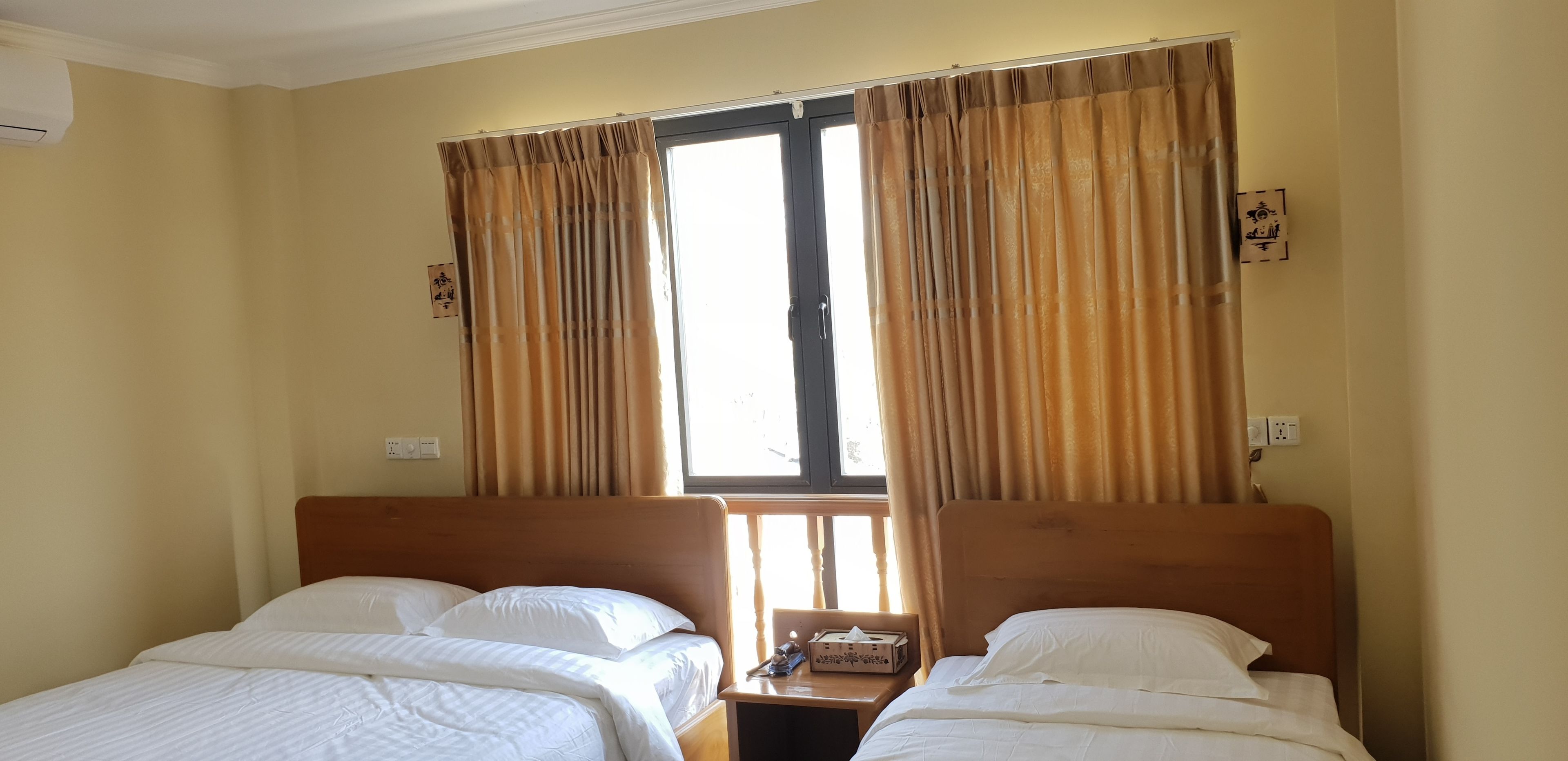 A hotel room with two single beds, beige walls, a window with curtains, and a bedside table with a lamp and a box.