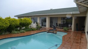 A swimming pool with a tiled deck, surrounded by greenery and a covered patio area. - Shelbourne Sea (Port Shepstone)