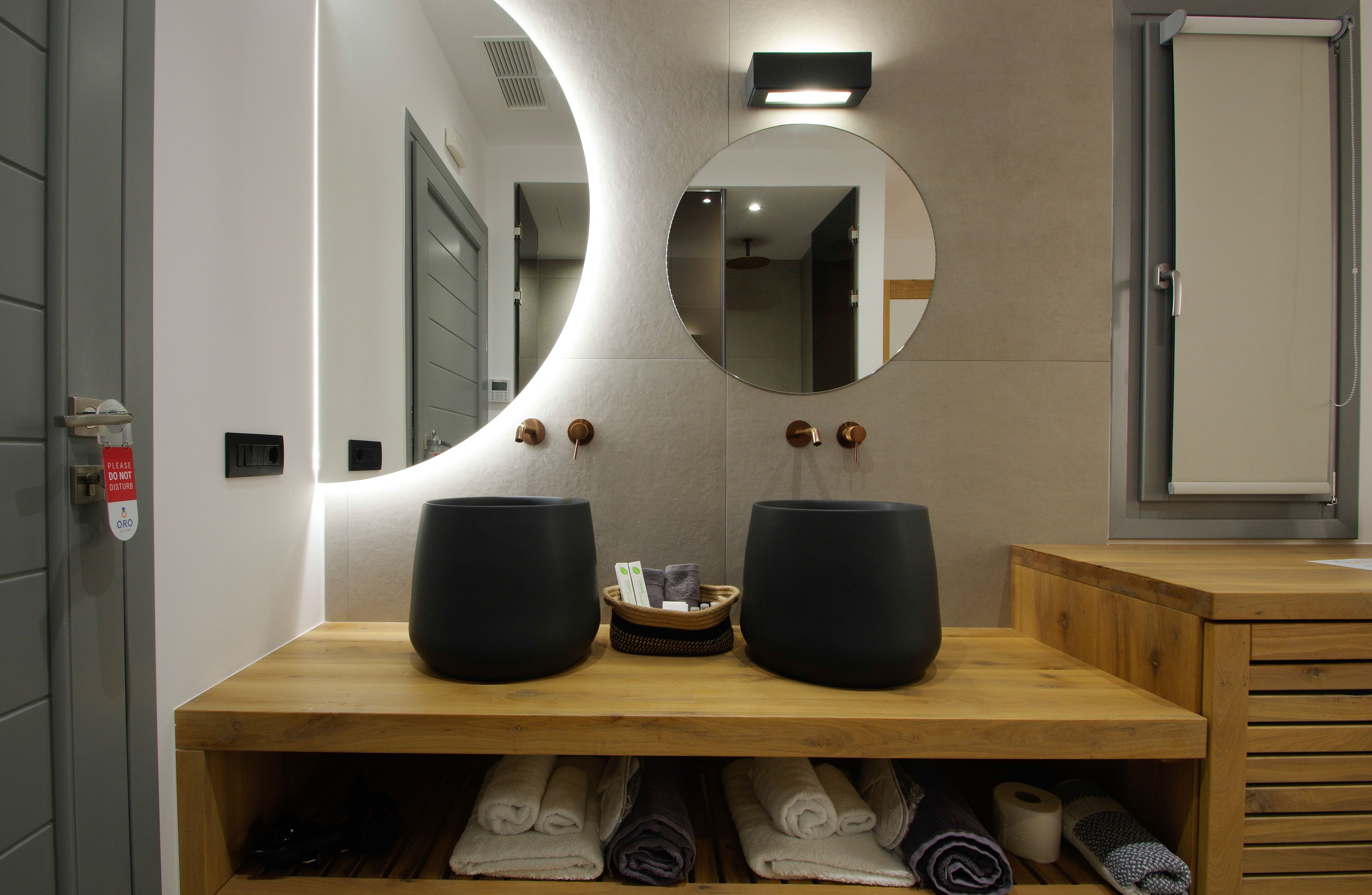 Un baño moderno con un mueble de tocador de madera, dos lavamanos negros, un espejo redondo y un perchero con toallas dobladas con cuidado.