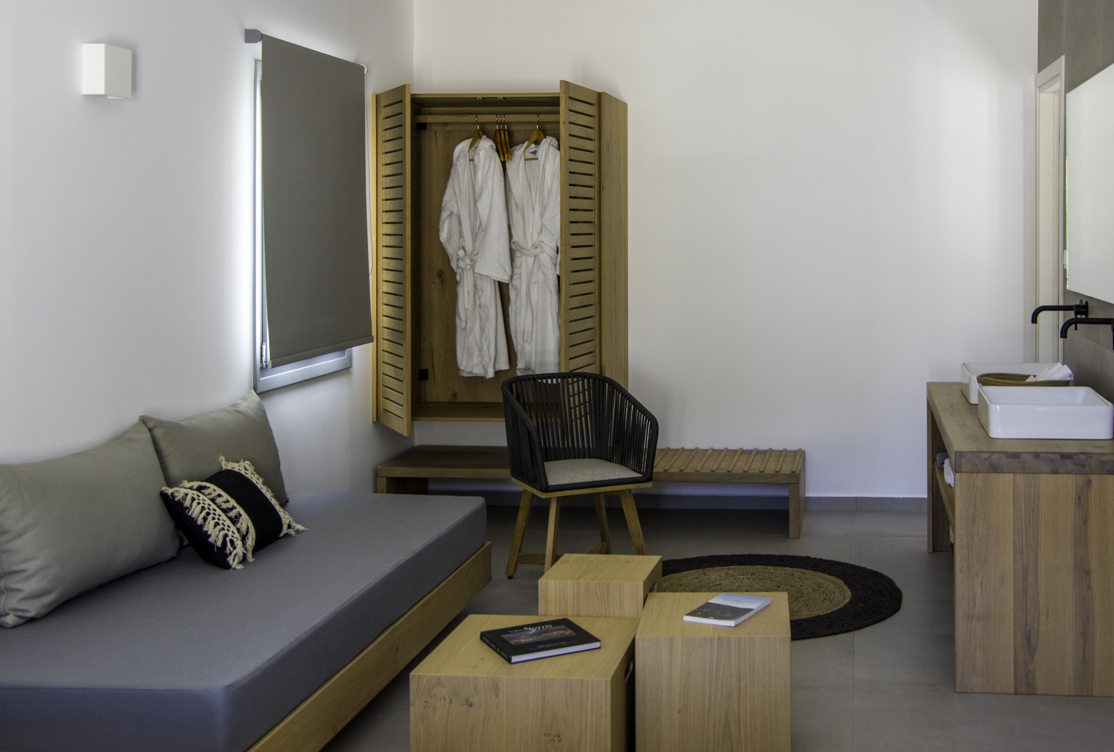 A modern room with a grey sofa, wooden side table, a black chair, a wooden bench, and a wooden wardrobe with clothes hanging.