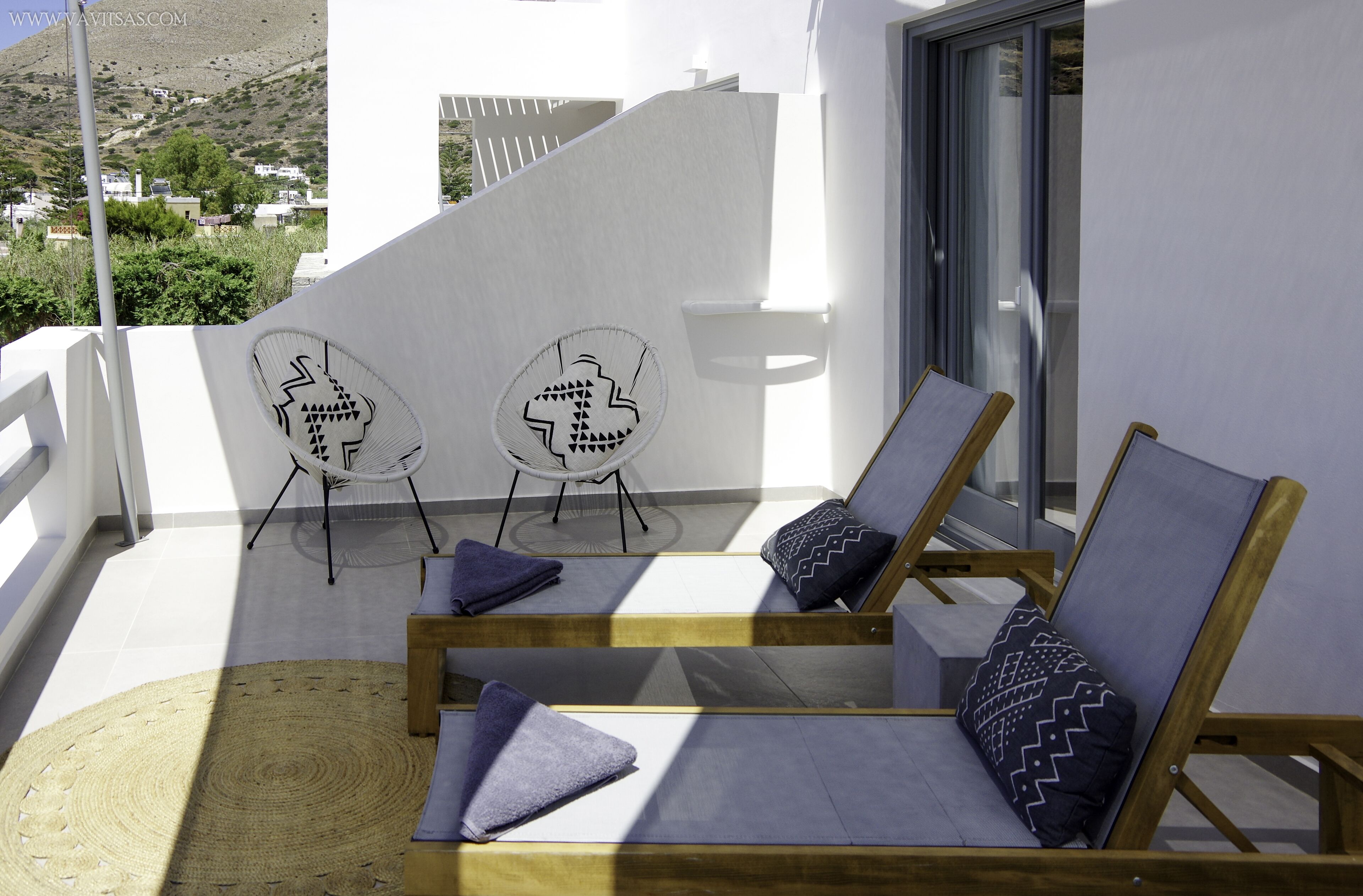 A balcony with two lounge chairs and a small table, overlooking a hillside.
