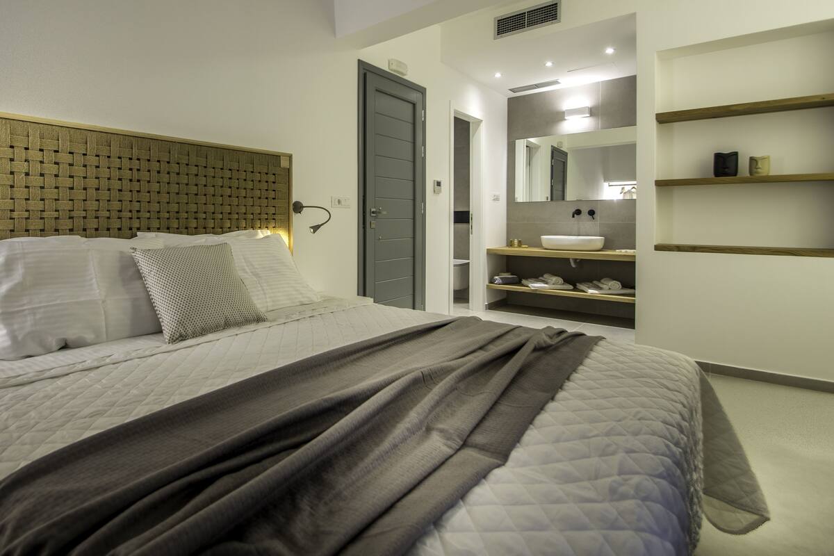 A modern bedroom with a large bed, a headboard with a woven pattern, a bedside table with a lamp, and a bathroom with a sink and shelves.