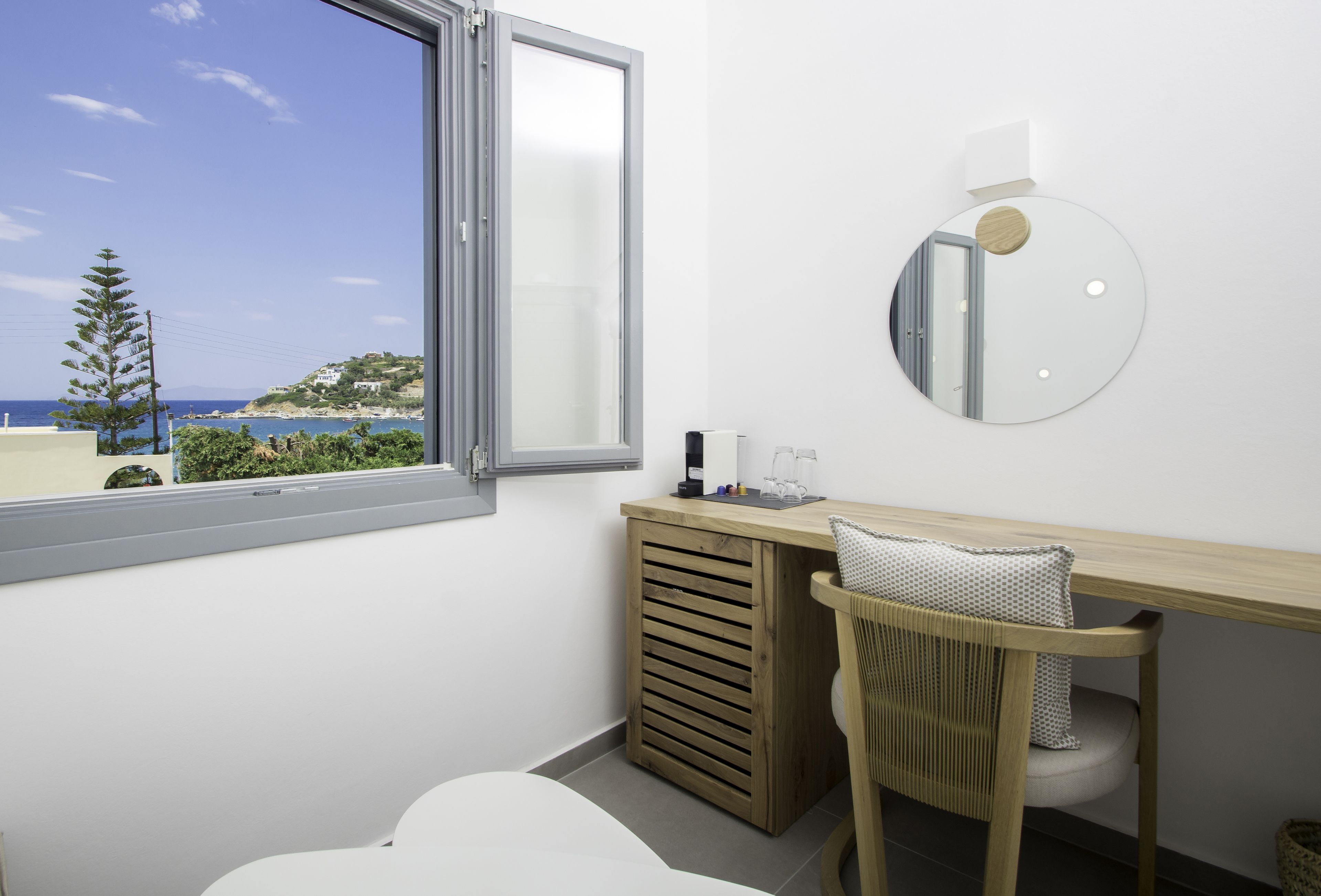 A modern bathroom with a large window, a round mirror, a wooden vanity, and a chair.
