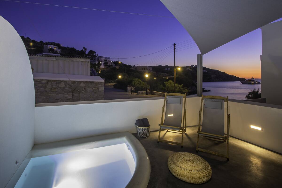 A balcony with a bathtub, two chairs, a small table, and a view of the sea at dusk.