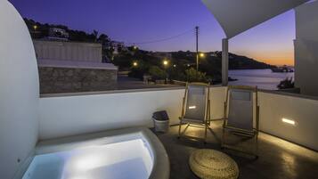 A balcony with a bathtub, two chairs, a small table, and a view of the sea at dusk.