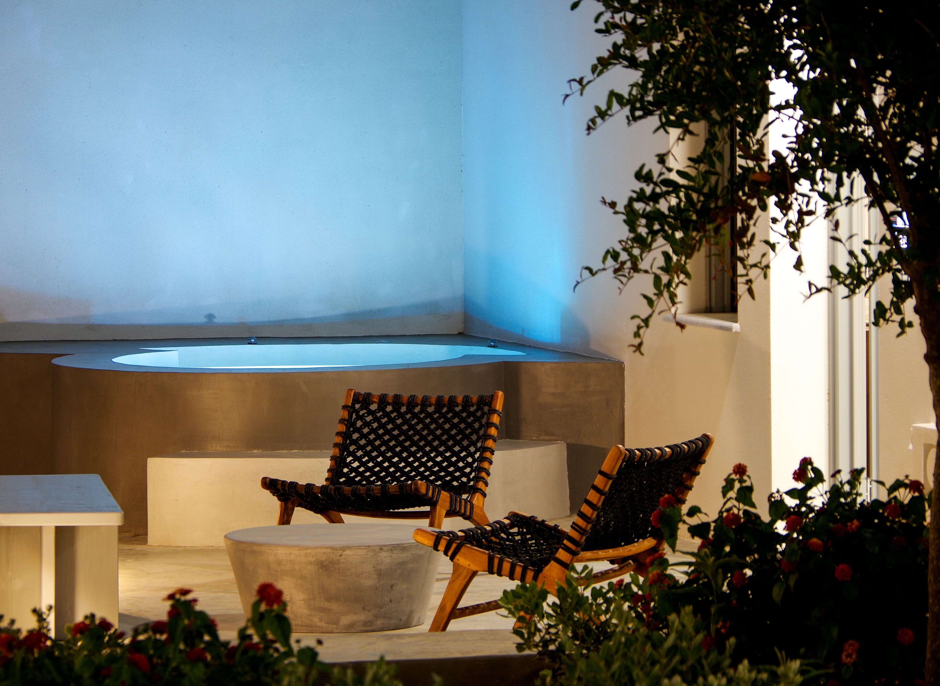 Un área de estar moderna al aire libre con dos sillas de mimbre y una pequeña mesa, un jacuzzi integrado y una jardinera de hormigón con flores.