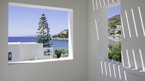 A room with a large window offering a view of the sea, a white wall, and a shadow pattern. - Oro Suites (Syros)