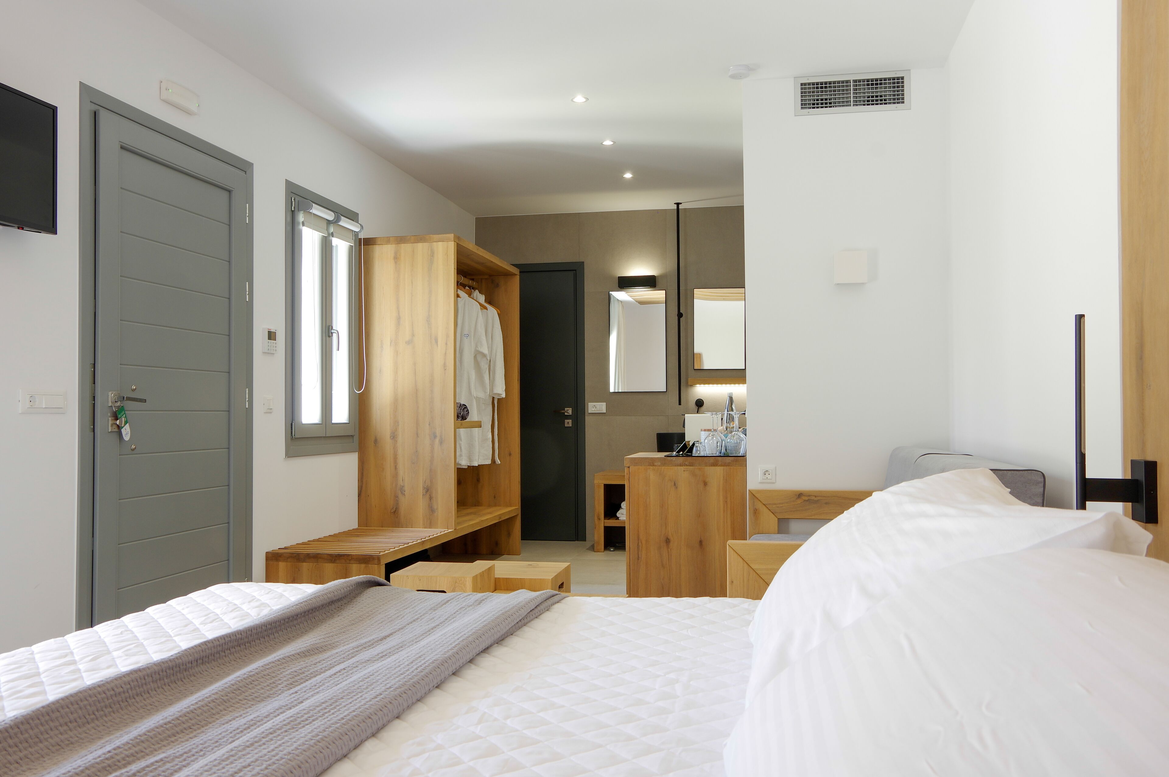A hotel room with a bed, a wooden wardrobe, a desk with a chair, a mirror, and a small bathroom area.