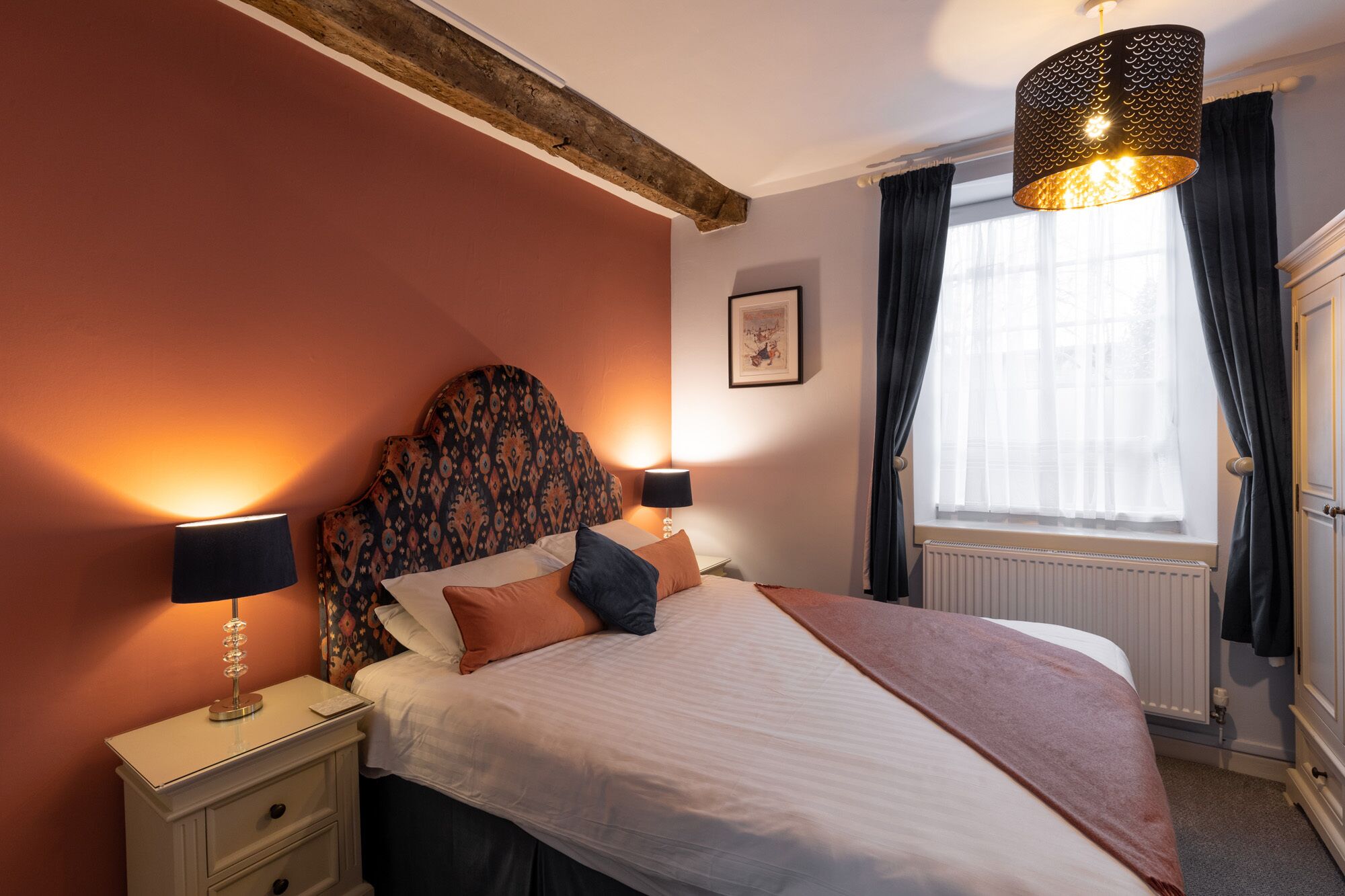 A bedroom with a patterned headboard, two bedside lamps, a bed with multiple pillows, a window with curtains, and a radiator.