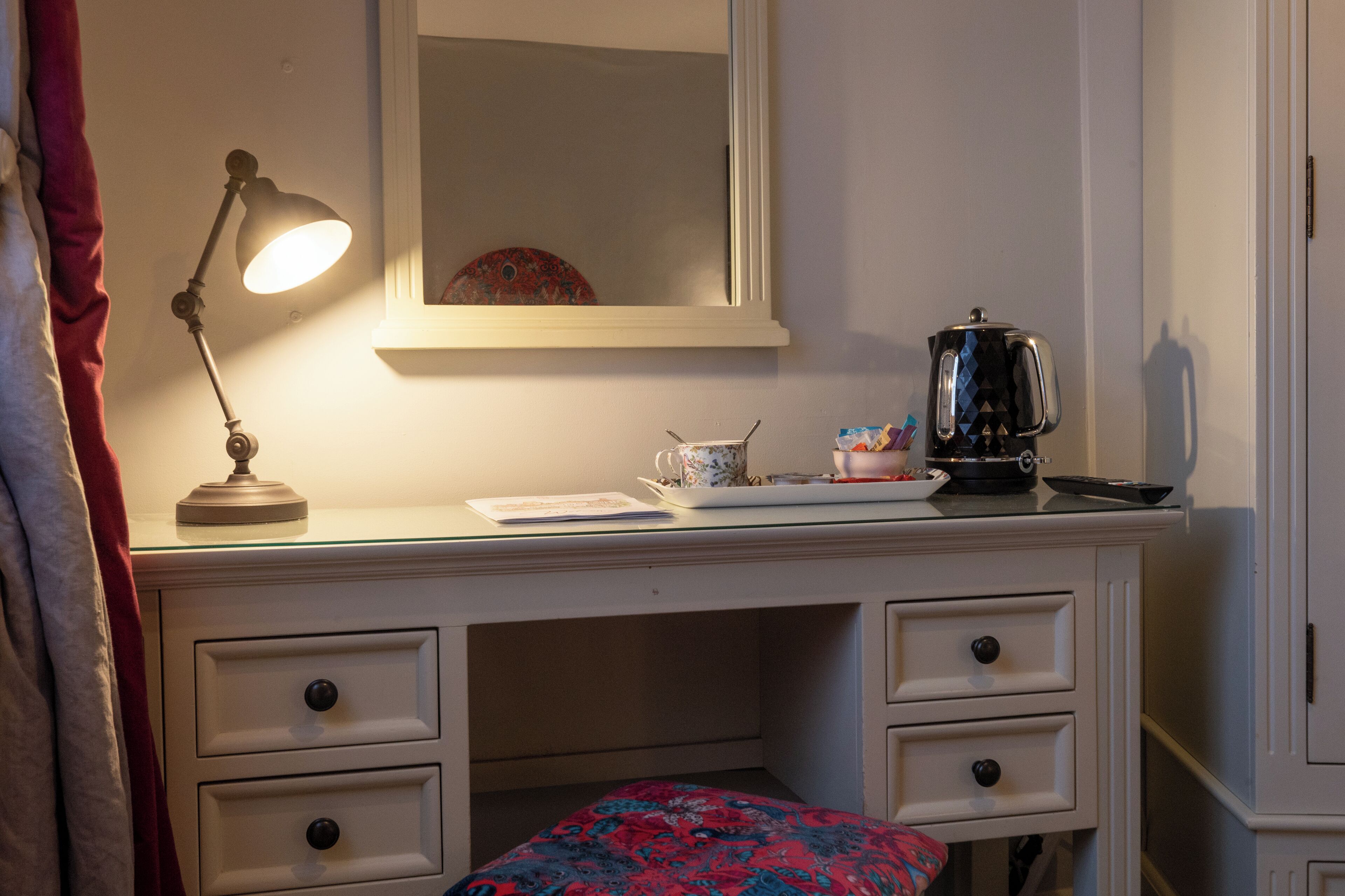 A white desk with drawers, a lamp, a mirror, a teapot, and a cup.