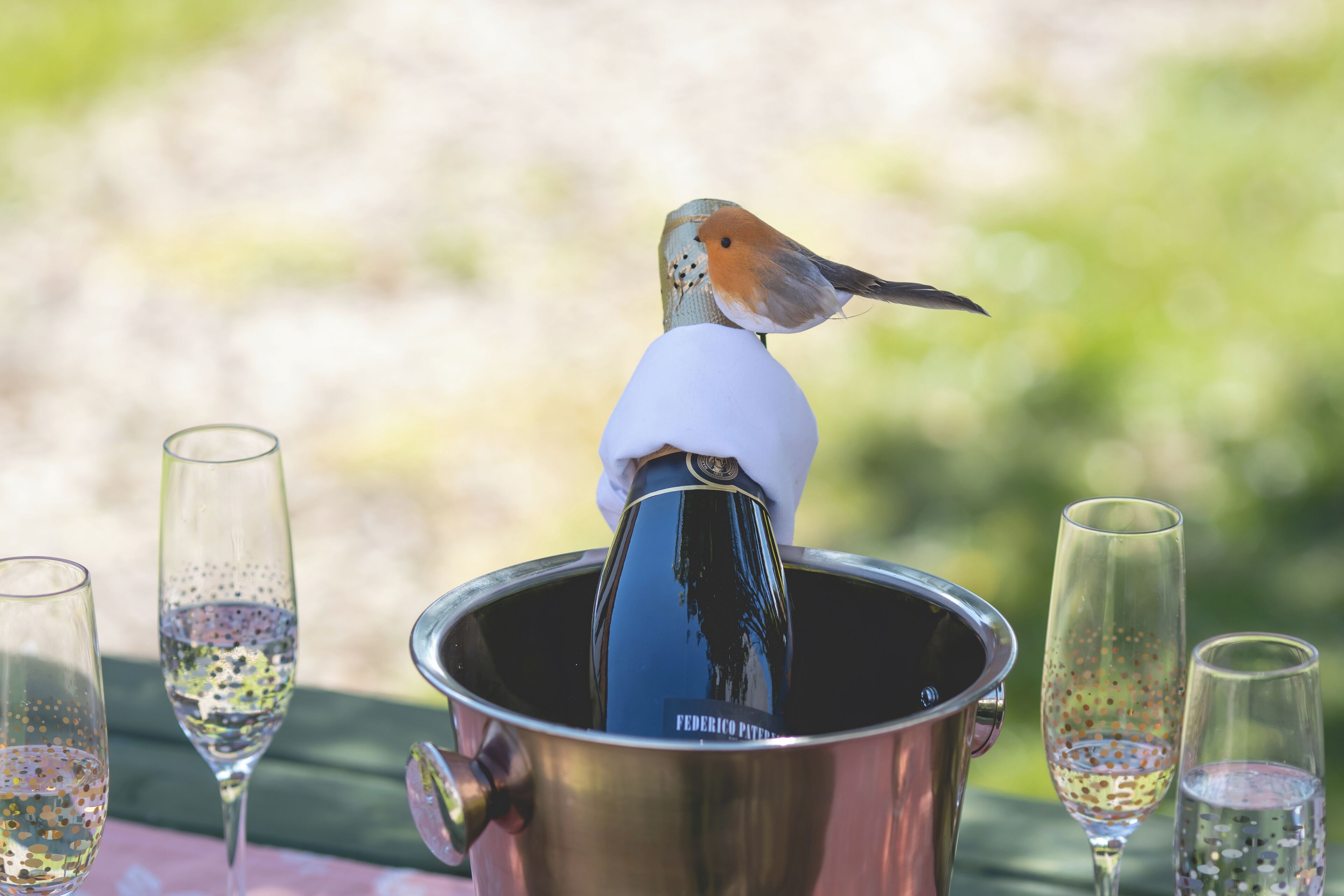 A bird perched on a white object above a metallic container with a label, surrounded by glasses with bubbles, on an outdoor table.