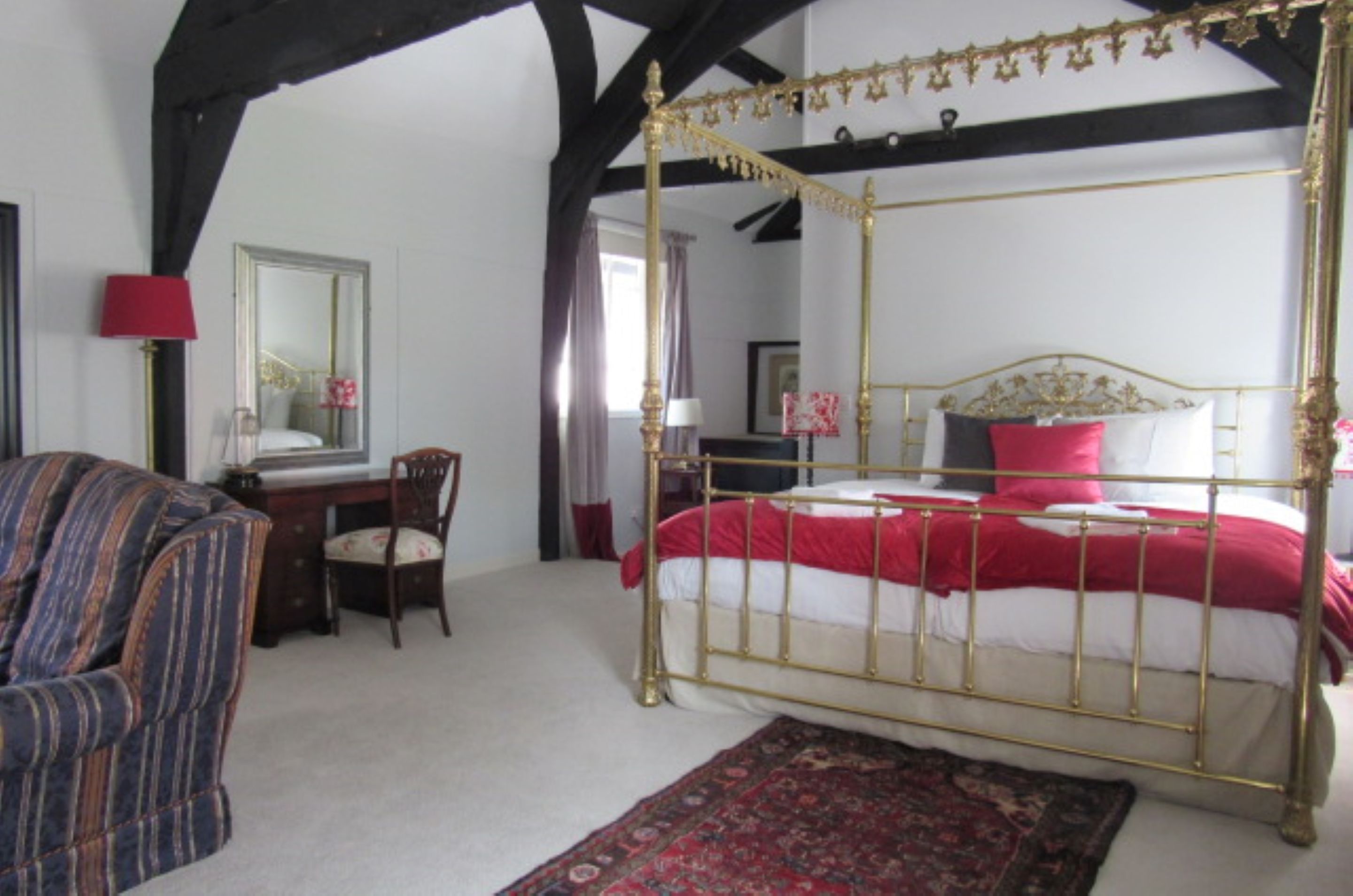 Een hemelbed met een rode en witte spreideken, een houten stoel, een bureau met een lamp en een spiegel aan de muur.