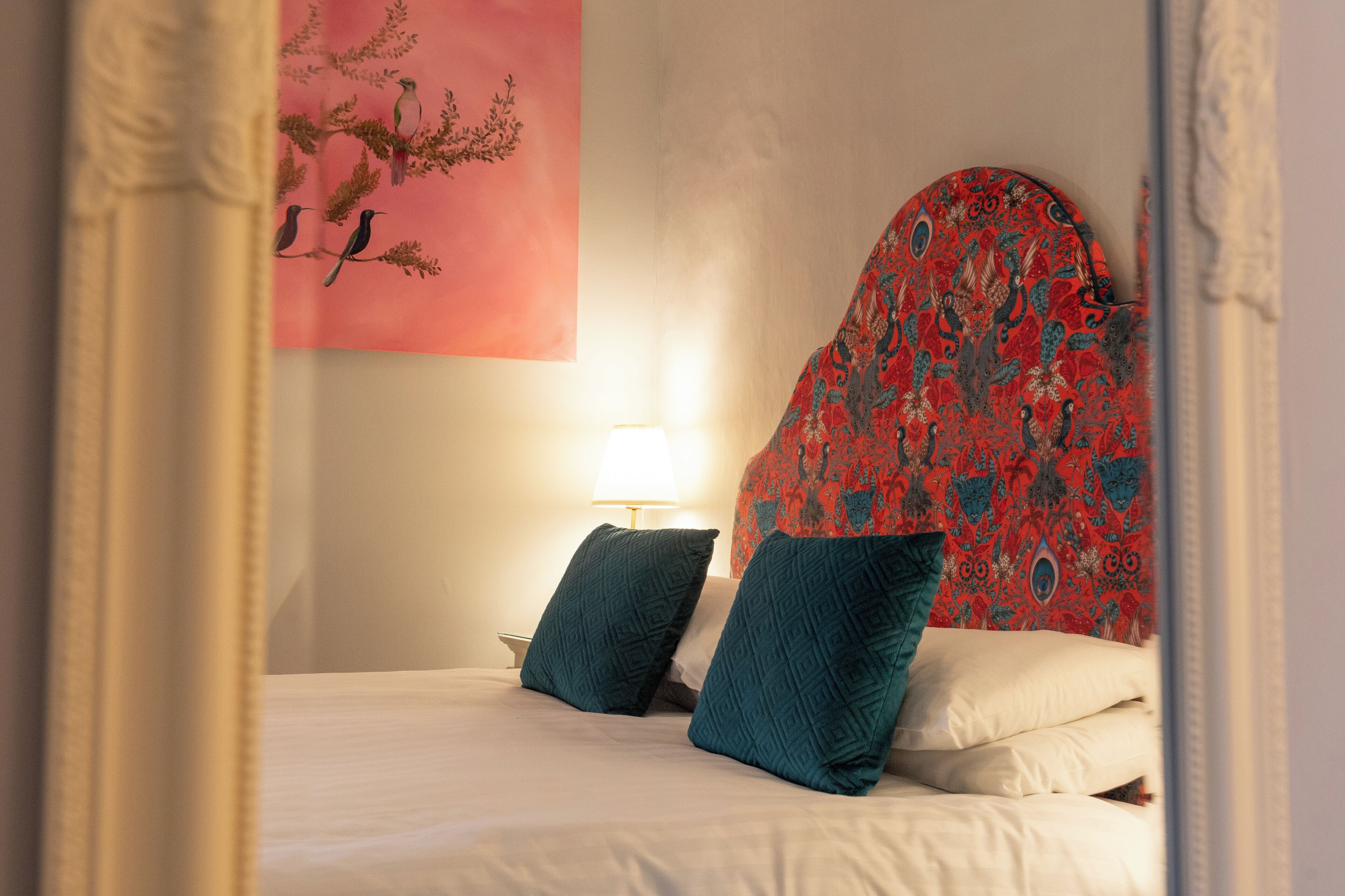 Cama com almofadas azul-turquesa, cabeceira estampada em vermelho e quadro de pássaros na parede cor-de-rosa.