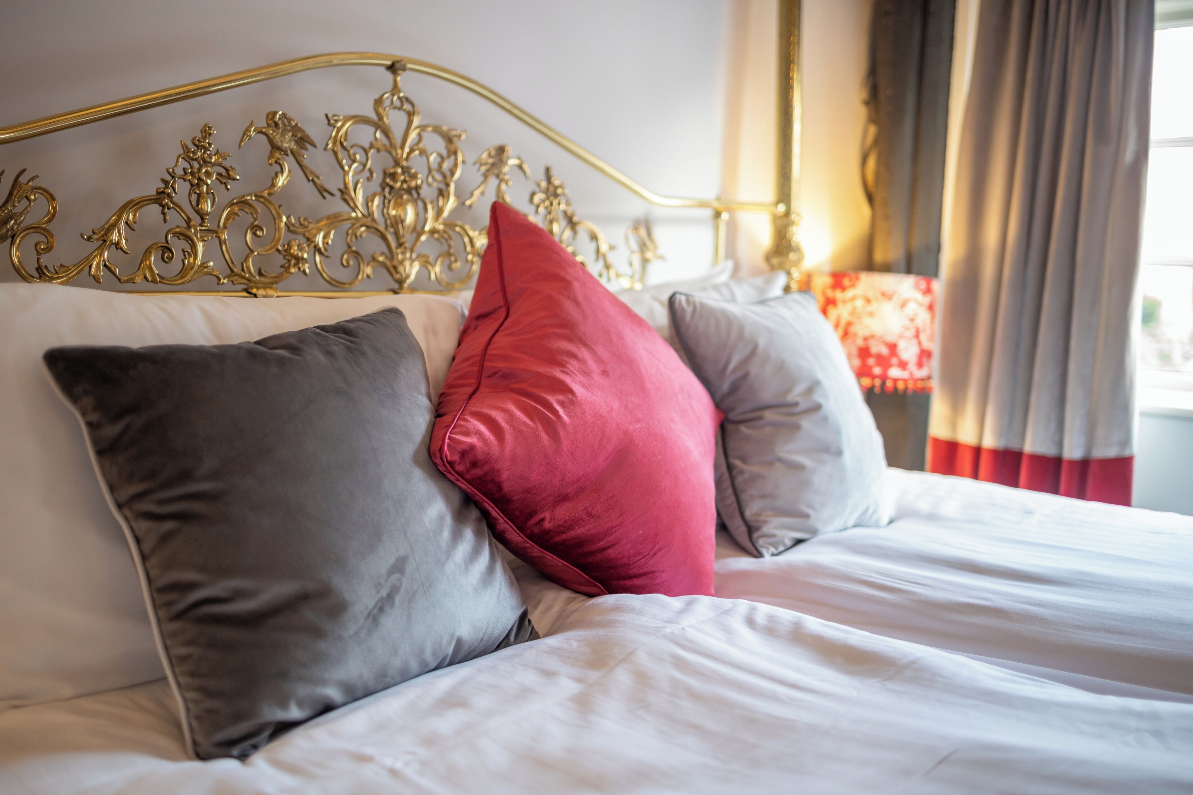 A bed with a gold headboard, white bedding, and multiple pillows in shades of gray and red.
