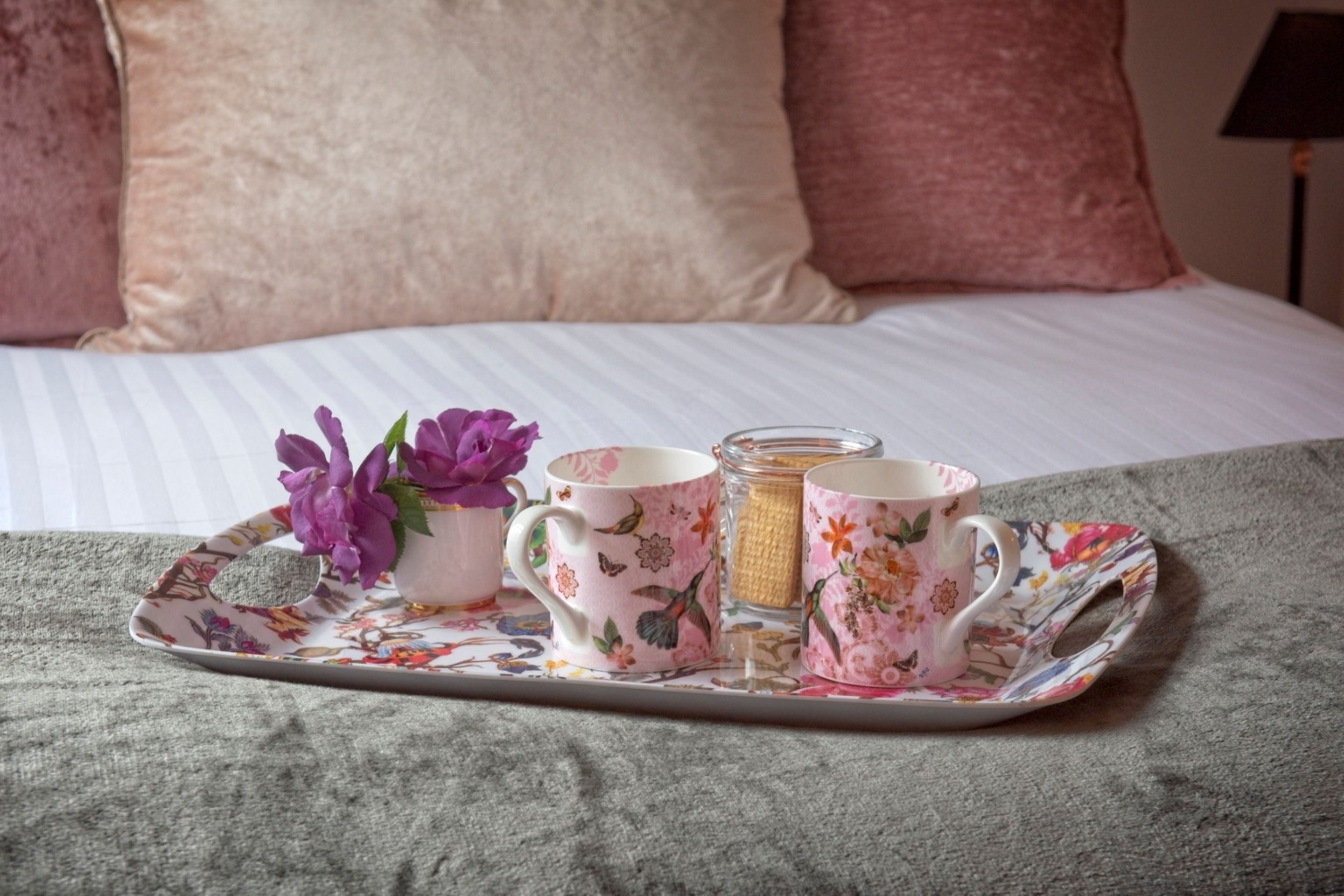 Una bandeja con tazas de estampado floral y un vaso sobre una cama con ropa de cama a rayas y almohadas rosas.