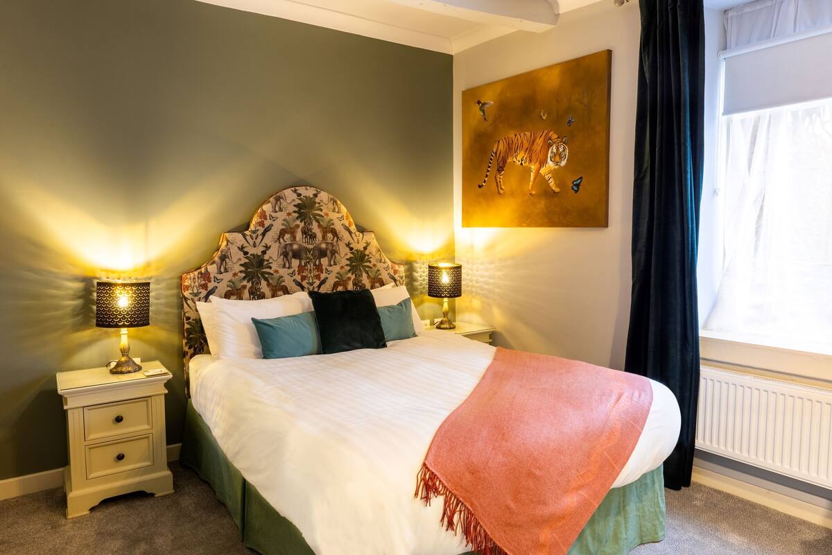 Un lit bien fait, agrémenté d’une tête de lit à motifs floraux, de deux lampes de chevet et d’un grand tableau représentant un tigre, accroché au mur.