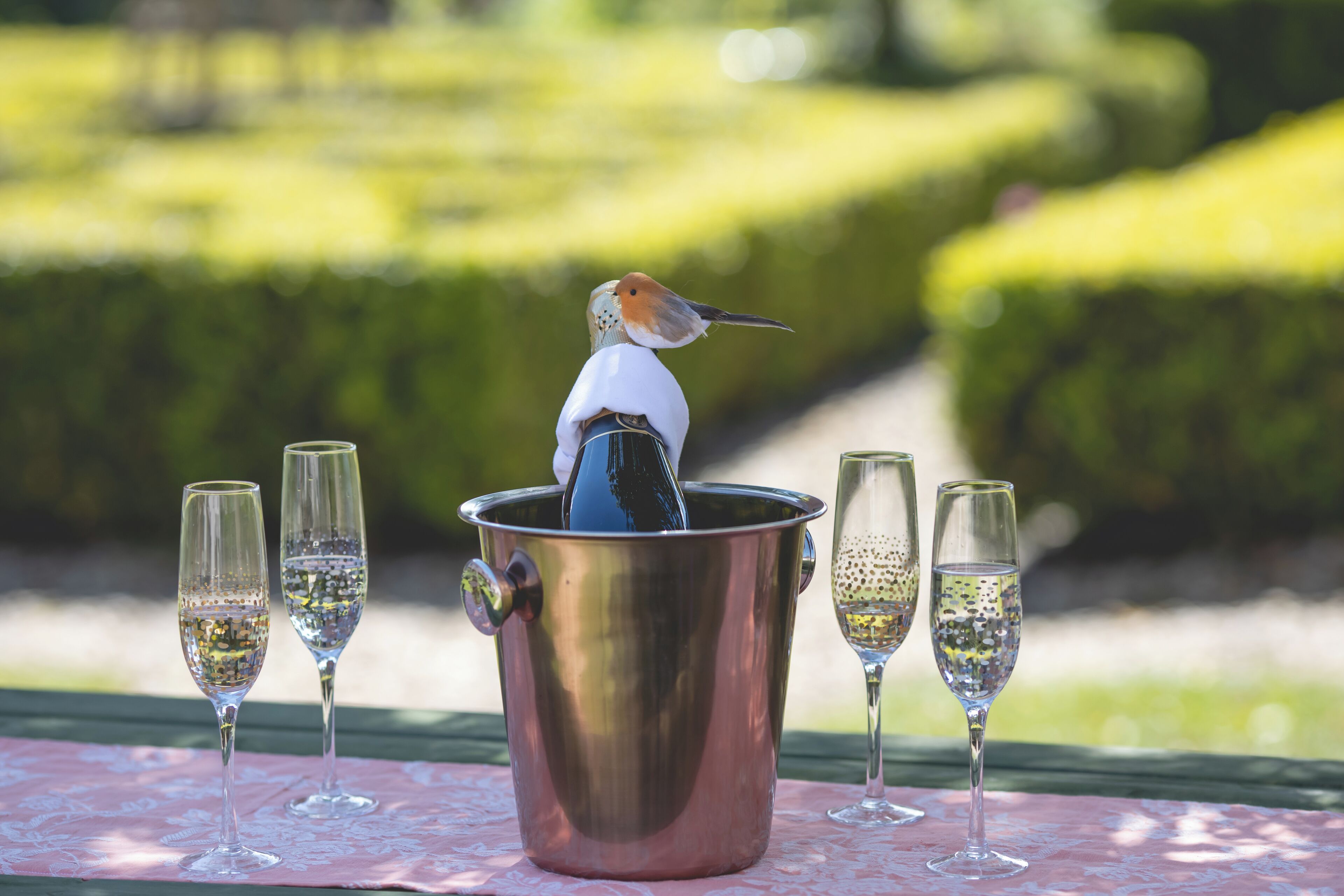 A bird in a metal container surrounded by champagne flutes on a table outdoors.