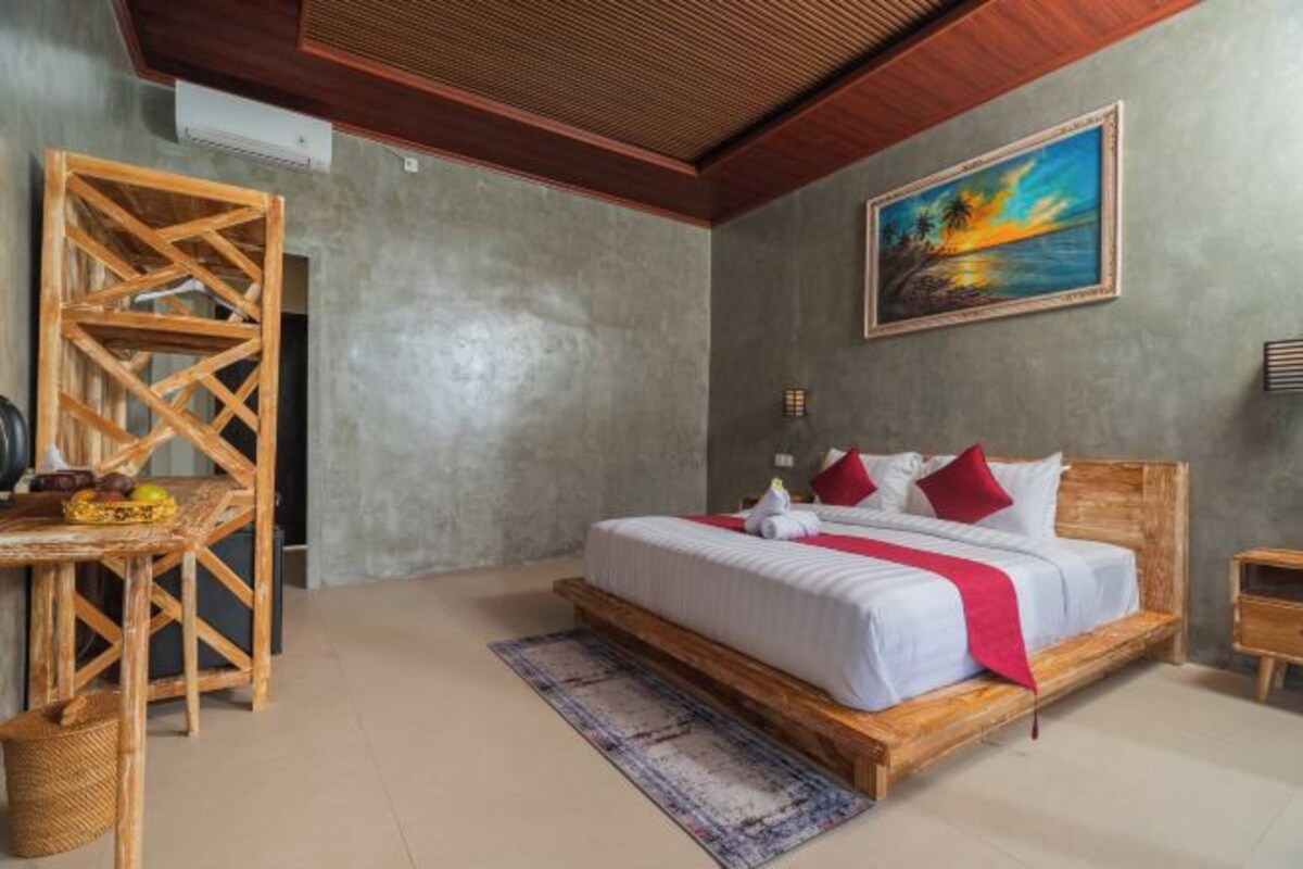 A bedroom with a wooden bed, a desk, a chair, a painting, and a view of the outdoors.