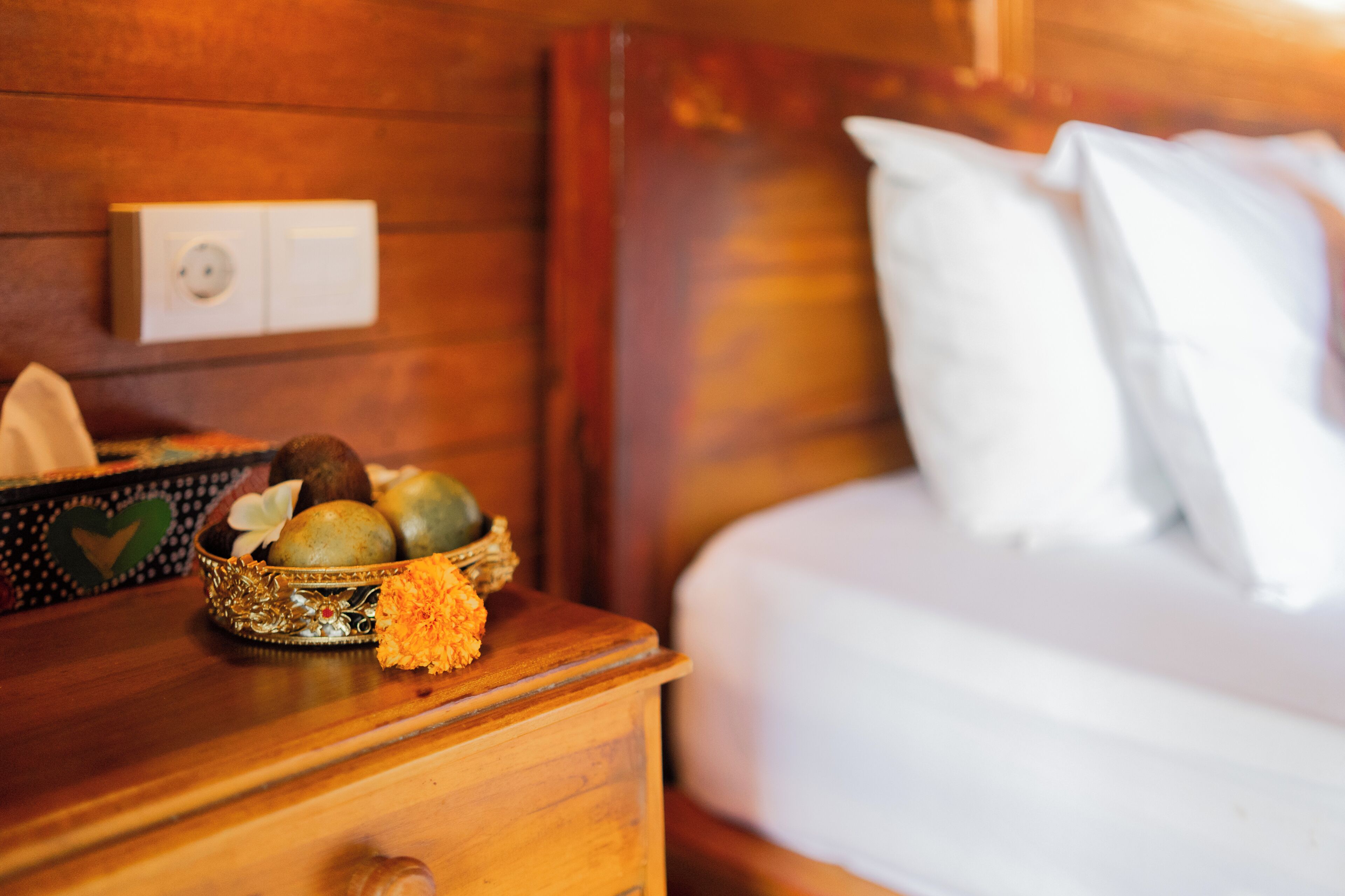 Un comodino in legno con una ciotola decorativa piena di frutta e fiori, un letto bianco con cuscini e un interruttore della luce fissato al muro.