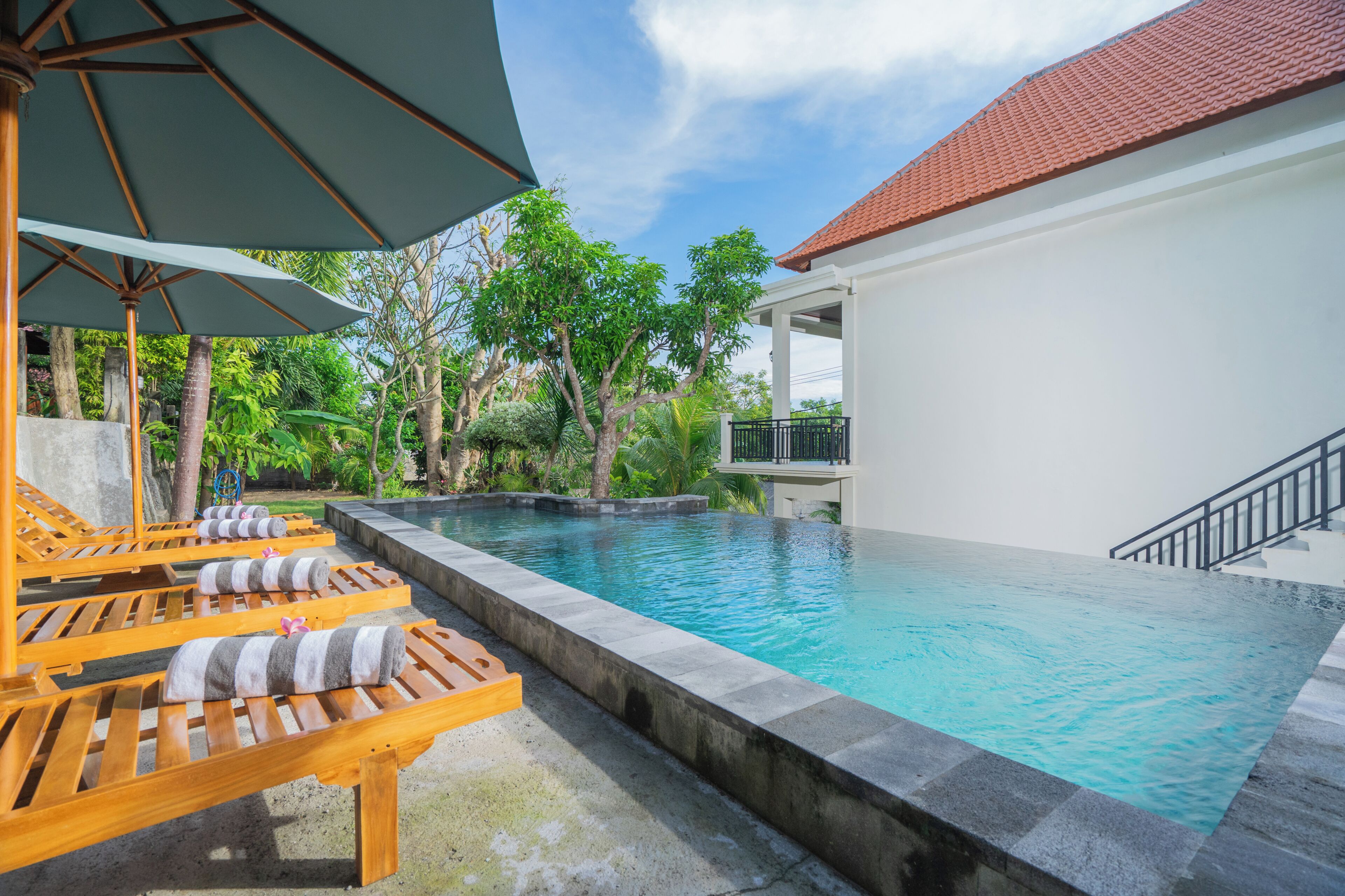 A poolside area with lounge chairs and umbrellas, surrounded by greenery and a building with a tiled roof.