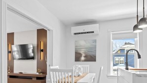 Une cuisine moderne avec des meubles blancs, un coin repas comprenant une table et des chaises blanches, un téléviseur à écran plat fixé au mur et une fenêtre donnant sur une maison.