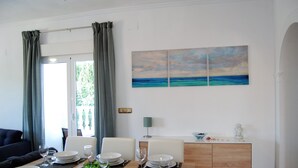 Un comedor con vista al mar a través de una ventana, un cuadro marino en la pared y un mueble blanco con una lámpara.