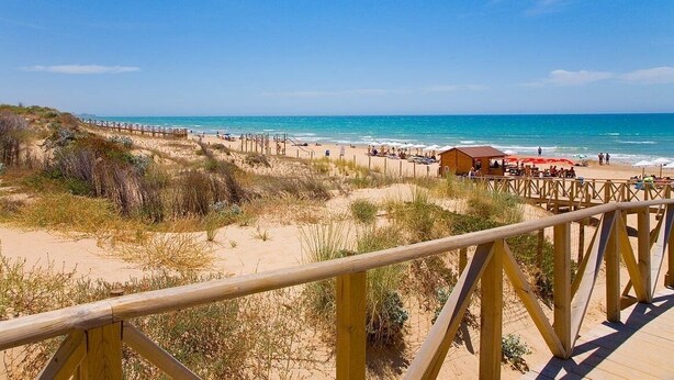 Una playa con un paseo marítimo de madera, dunas de arena y una estructura de madera con sombrillas.