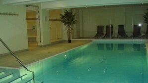 Una piscina interior con agua cristalina, un conjunto de escaleras que conducen a la piscina y varias sillas de descanso junto a la pared.