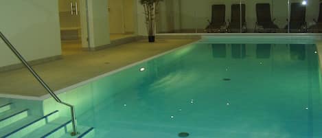 Una piscina interior con agua cristalina, un conjunto de escaleras que conducen a la piscina y varias sillas de descanso junto a la pared.