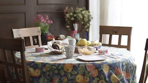 Una mesa de comedor con mantel estampado de frutas, un cuenco de fruta, una tetera y una taza.