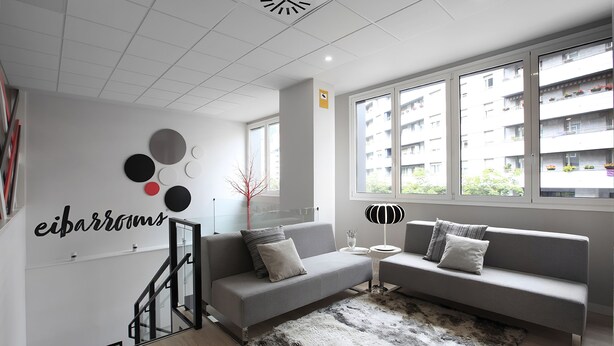 A modern living room with a grey sofa, a small side table, and a large window with a view of a building. - Eibarrooms (Eibar)